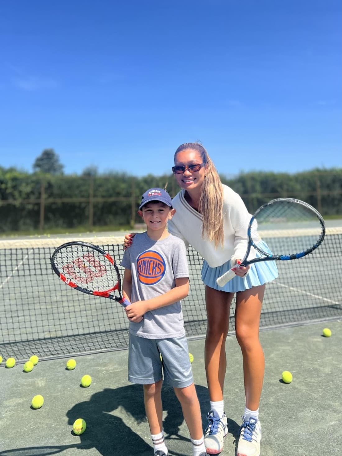 Bolor on a sunny court next to a young junior player, both holding rackets after a lesson.